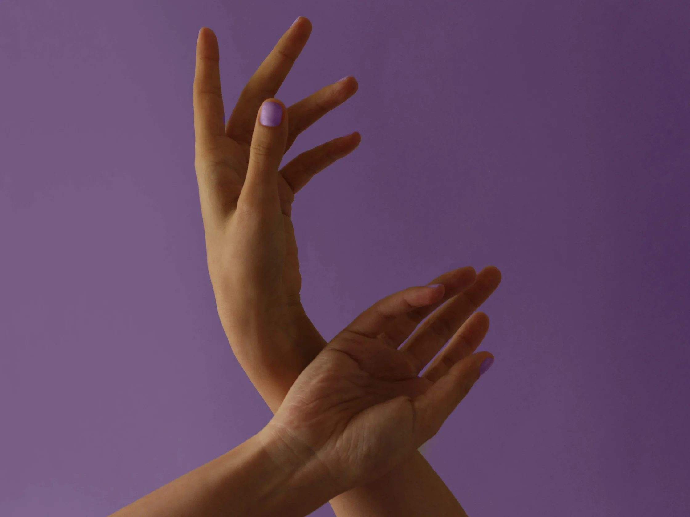Several hands intertwined against a solid purple background, nails painted lavender.
