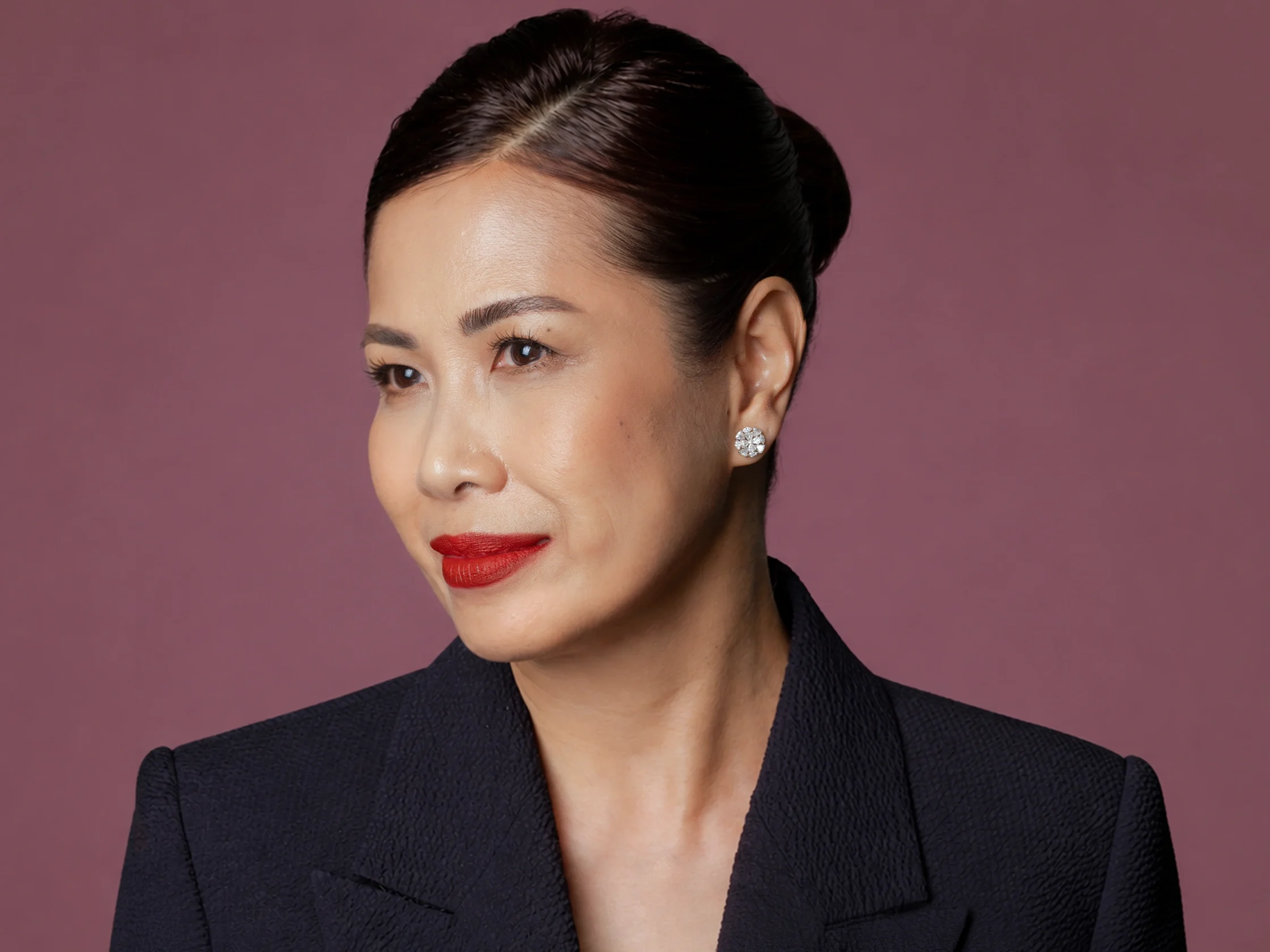 Close-up portrait of a woman with slicked-back hair, red lipstick, and diamond earrings against a muted maroon backdrop.