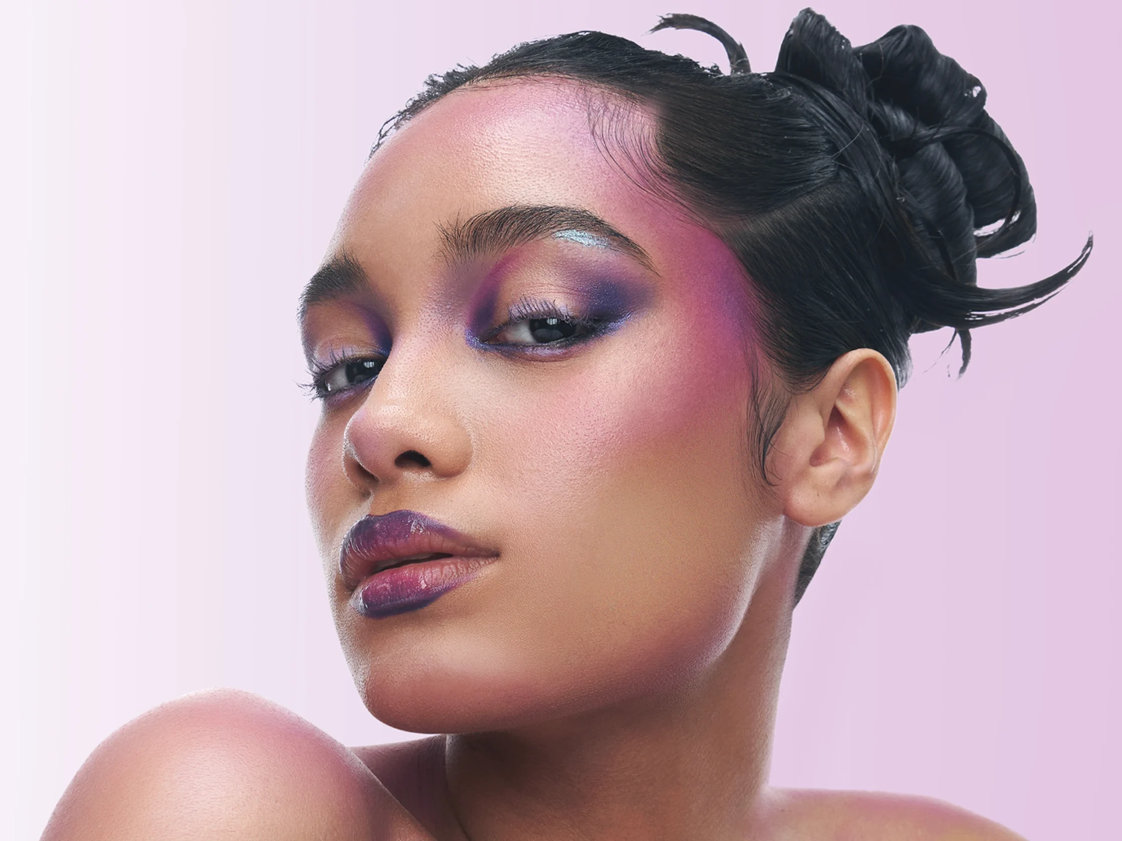 Close-up of a woman with bold pink and purple makeup against a pale pink background.