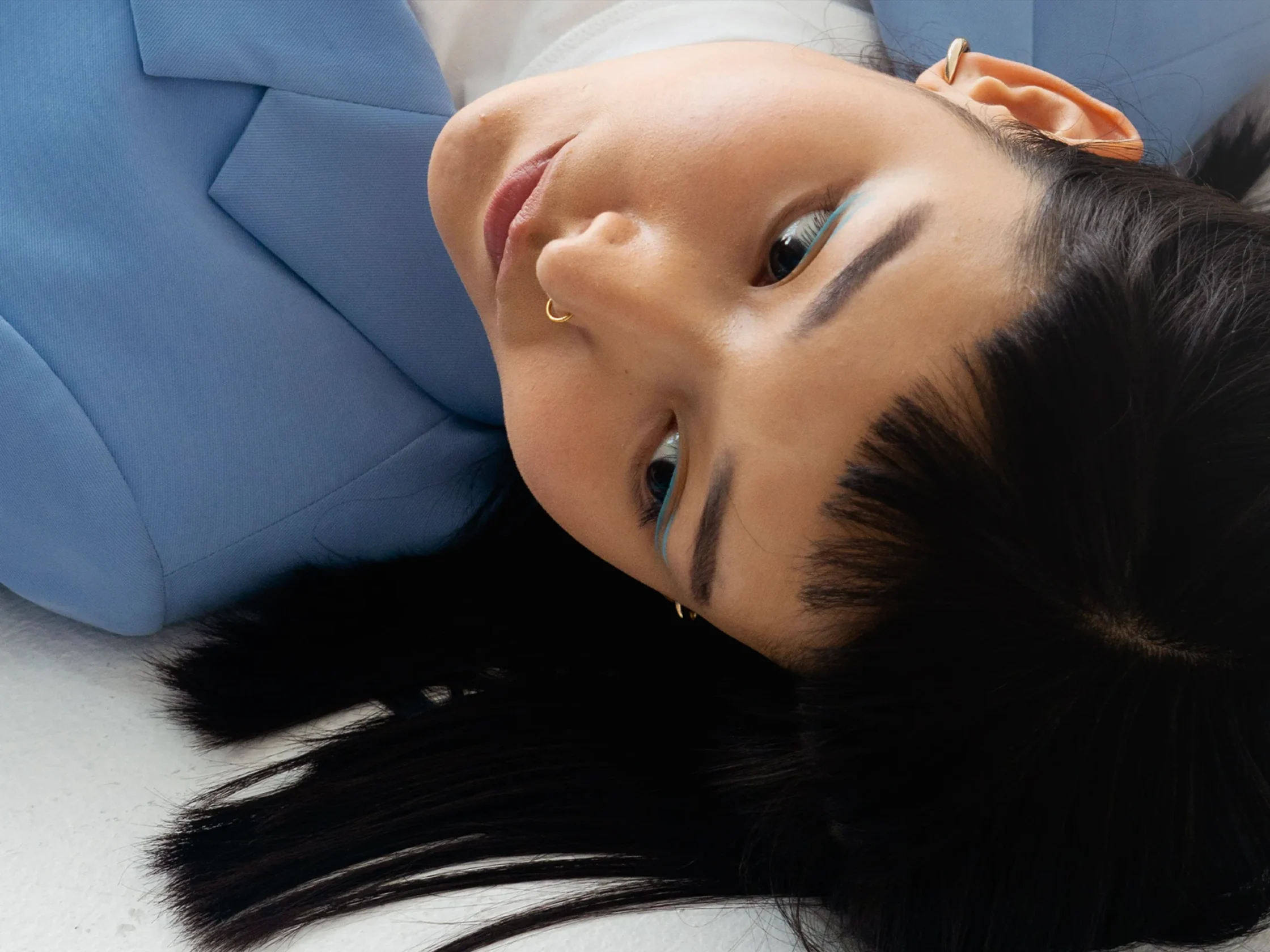 Close-up of a person lying on a blue cushion, wearing glasses and a small gold hoop earring, with long dark hair spread around.