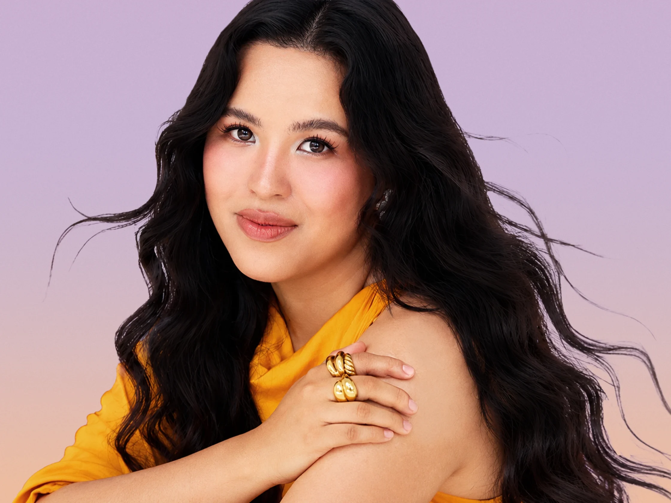 Close-up portrait of a woman with long dark hair and an orange top against a soft purple background.