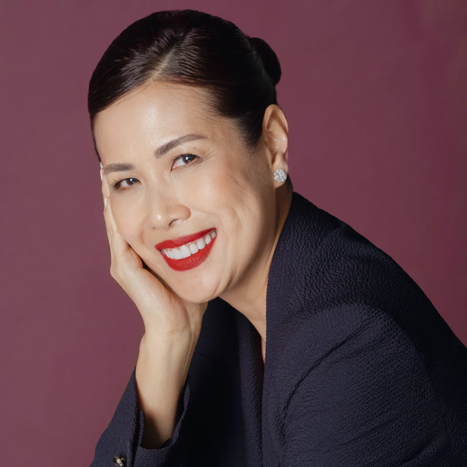 Smiling woman in a dark blazer with diamond stud earrings, viewed from the side against a maroon background.