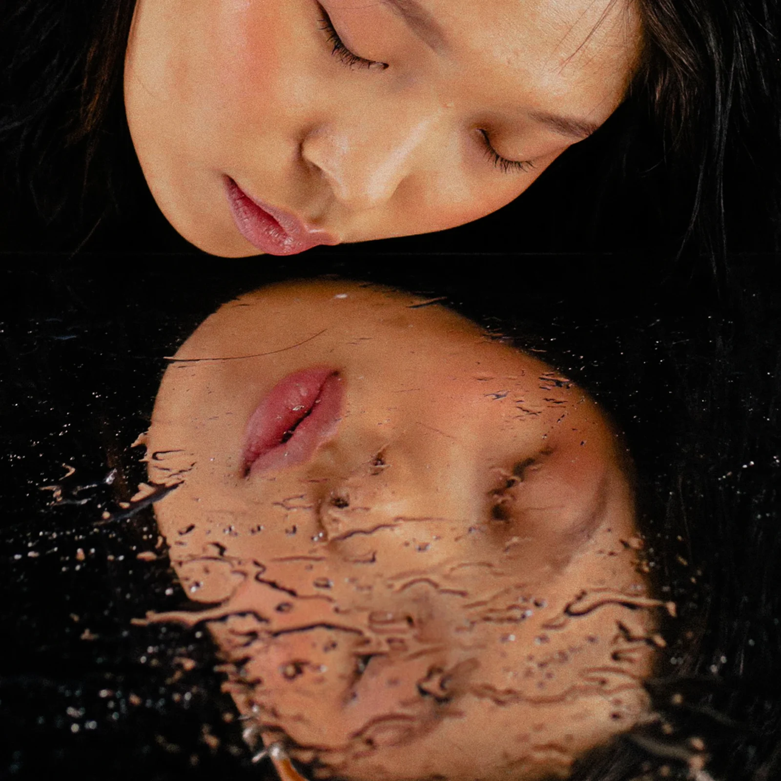 Close-up of a woman with eyes closed, leaning toward a dark surface with her reflection visible in the water/shine.
