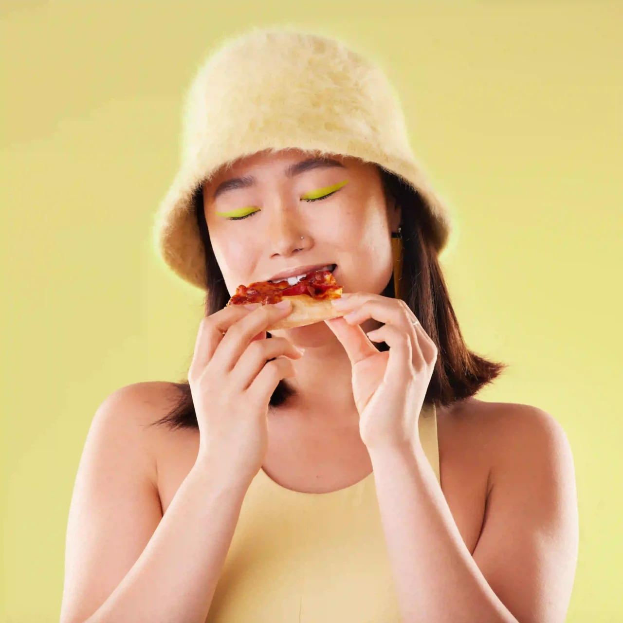 Woman wearing a fluffy beige hat, lime eyeliner, taking a bite of a pepperoni pizza against a pale yellow background.
