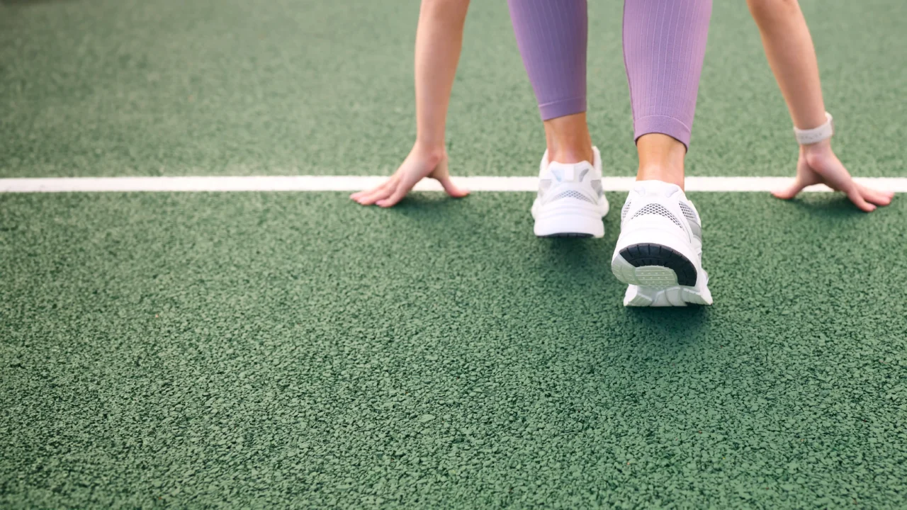 Runners in purple leggings ready at the starting line on a green track, wearing white sneakers, hands on the ground.