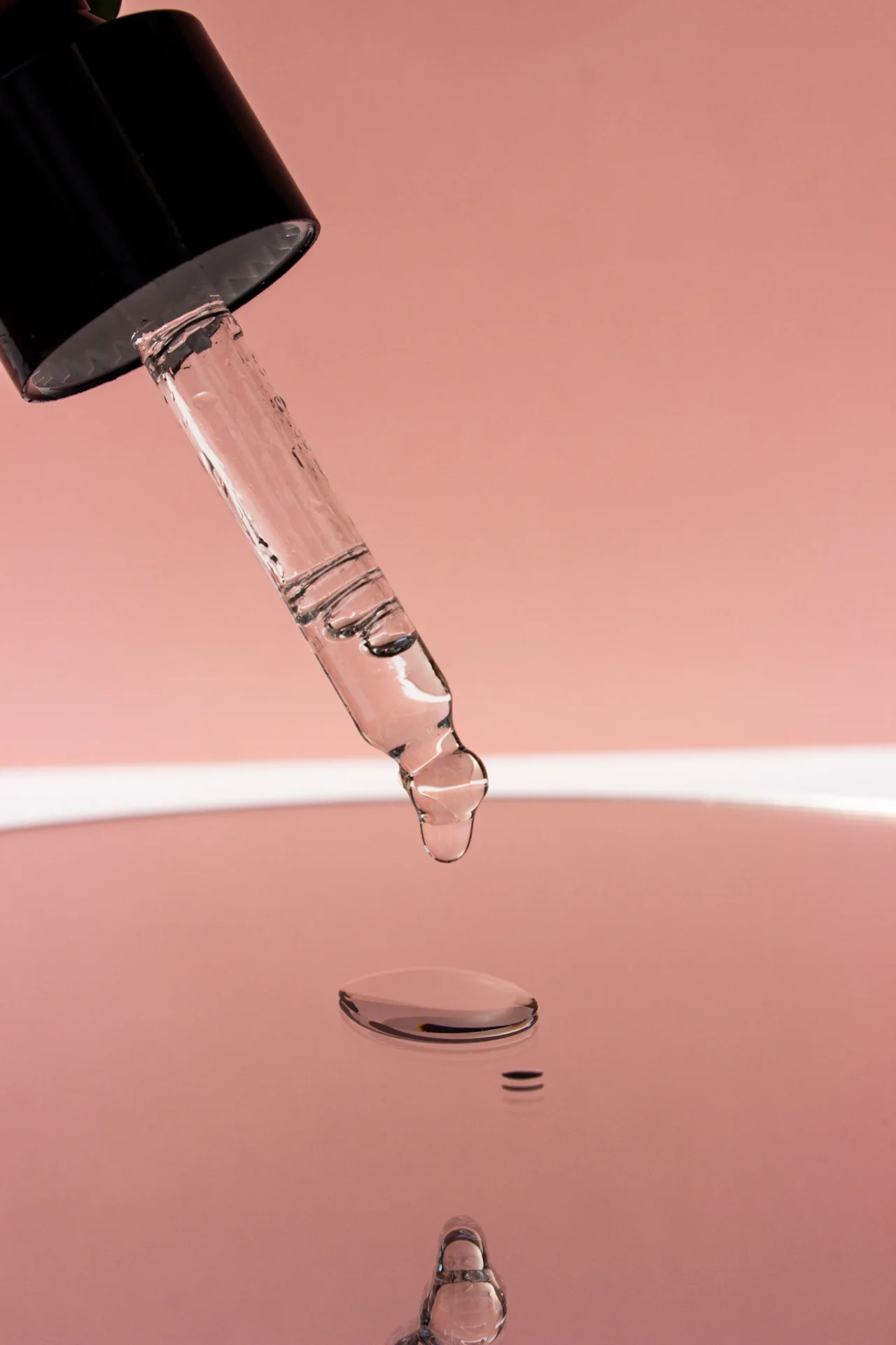 Close-up of a glass dropper releasing pink liquid against a pink background.