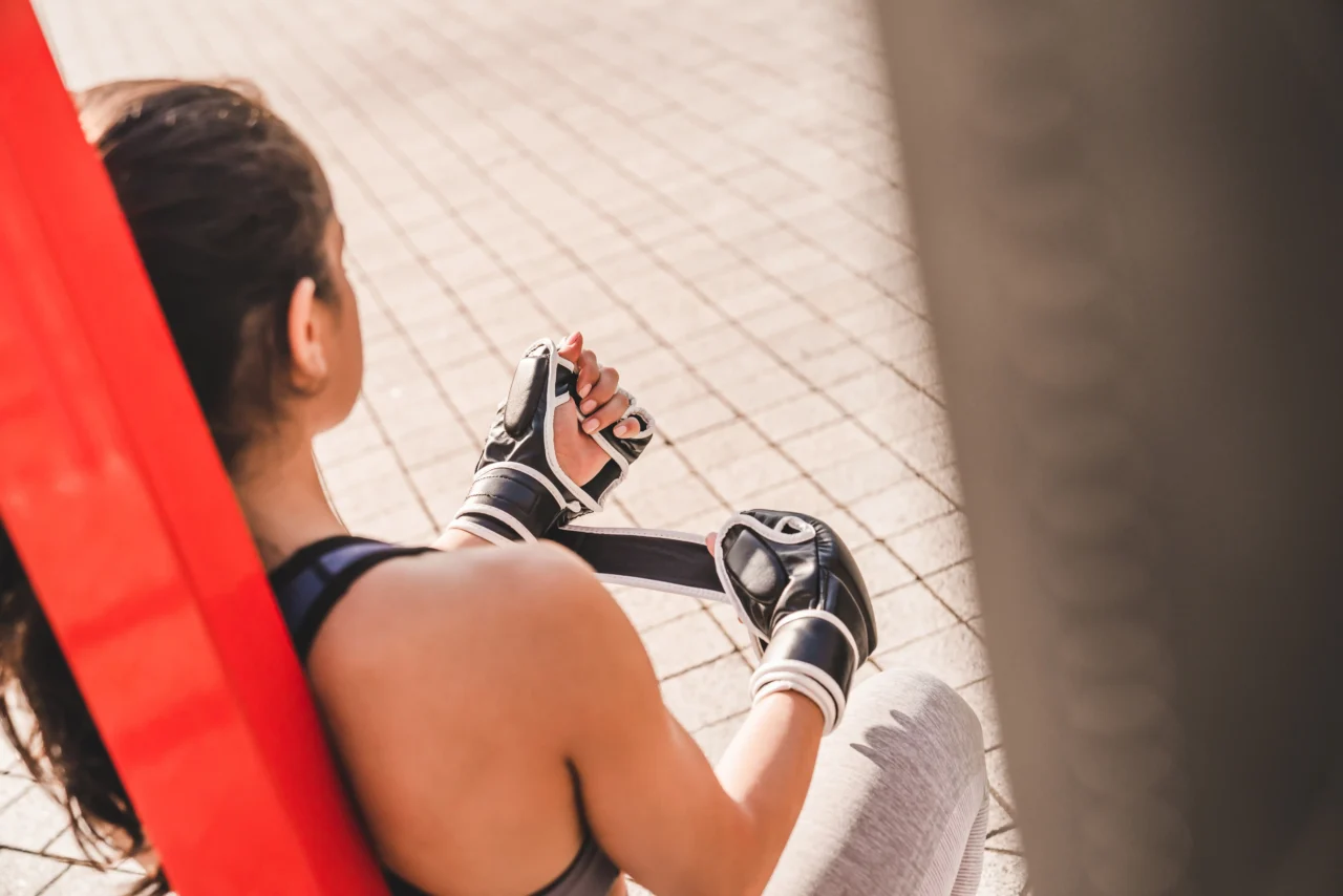 Person in a life jacket and gloves, gripping a strap while seated outdoors on a red surface to the left.